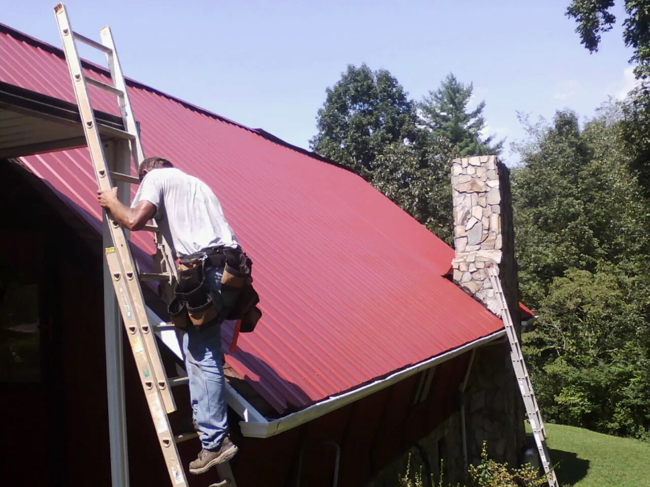 Roofer on a ladder beside a red metal roof and stone chimney — Soffit & Fascia Repair by Roofing Brothers in York Harbor